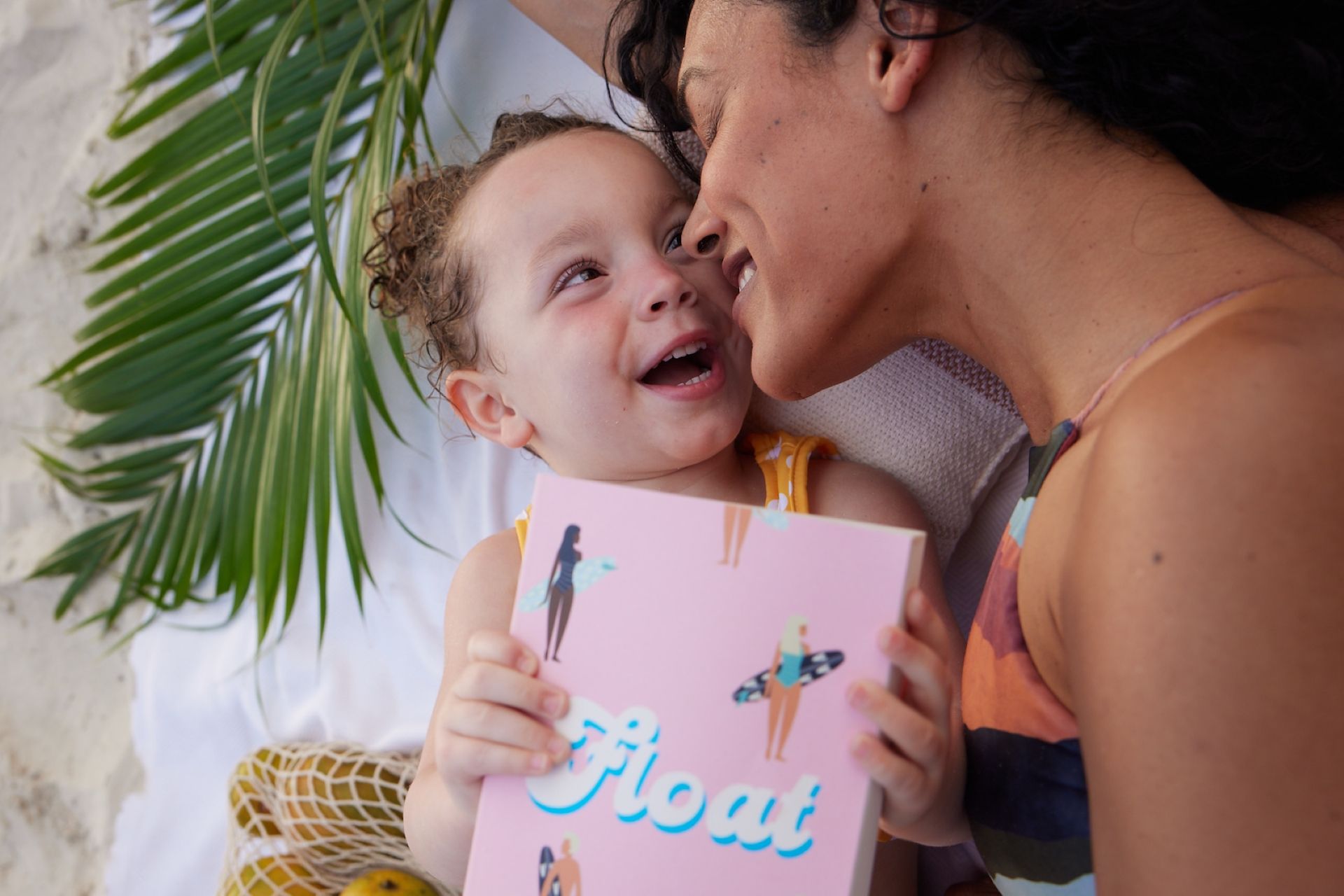 Smiling toddler holding a pink book titled "Float" while cuddling nose-to-nose with their mother under a palm leaf.