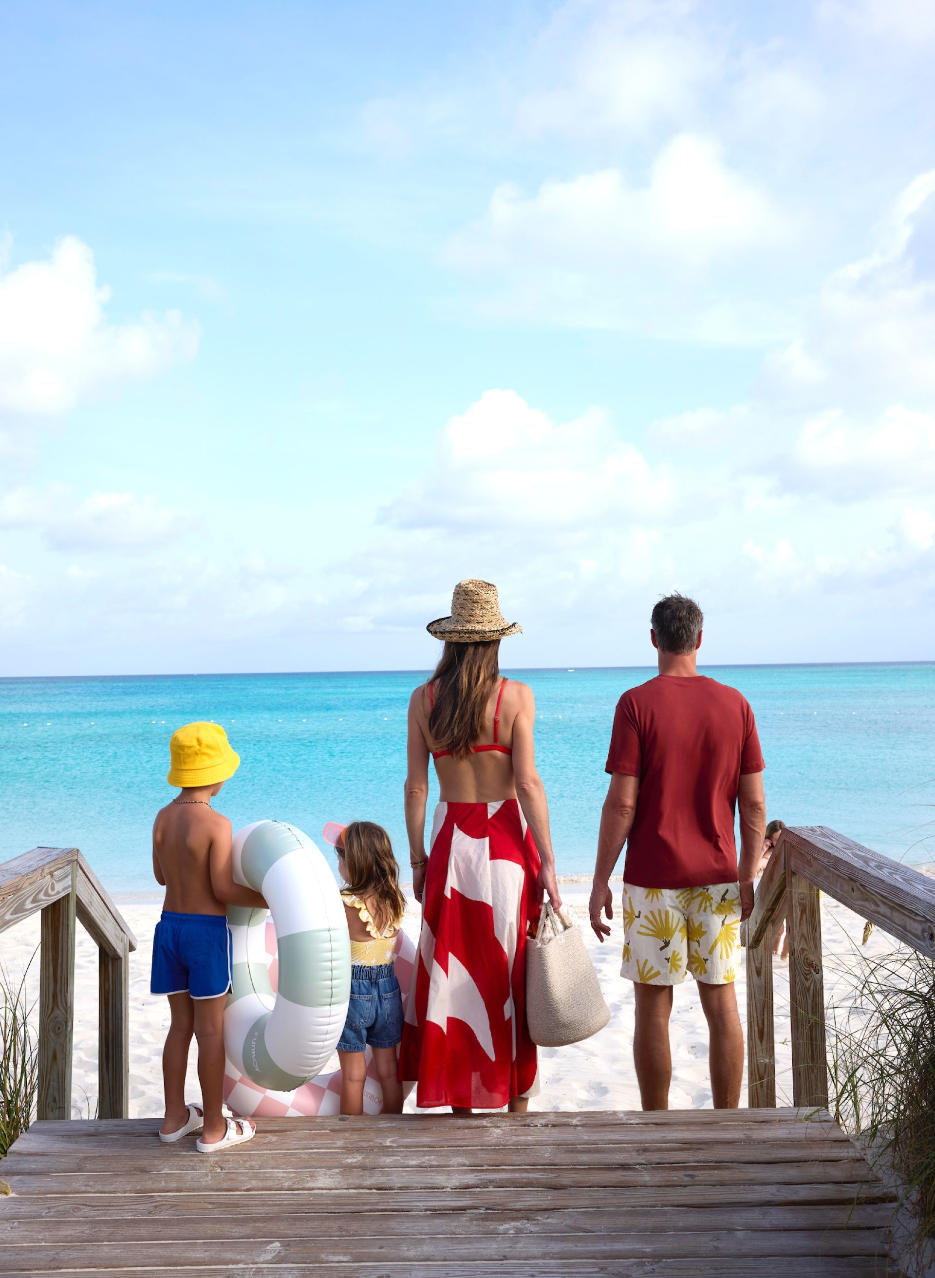 Family of four standing on a wooden walkway, looking out at the turquoise ocean, with beach gear and floaties in hand.