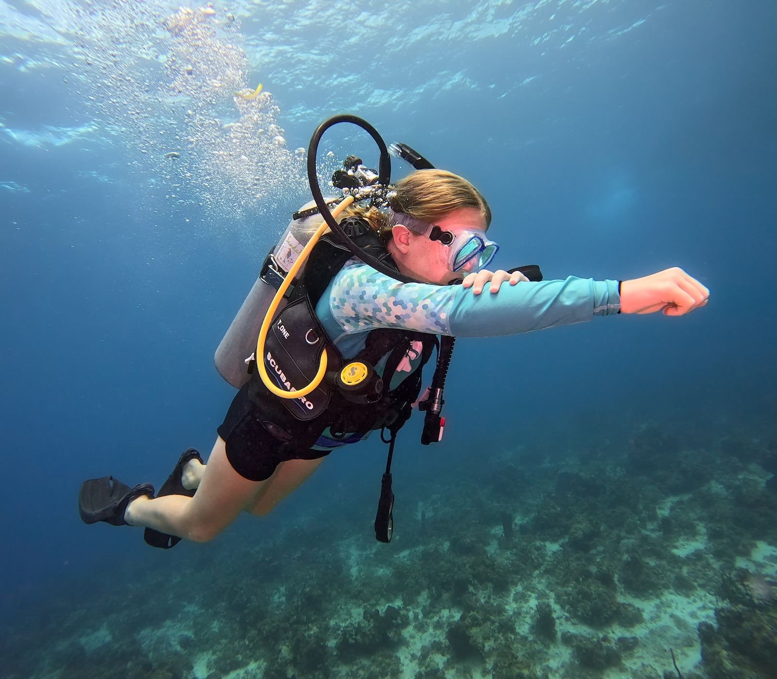 A child scuba diving underwater, wearing a mask and air tank, swimming above a coral reef in clear blue water.